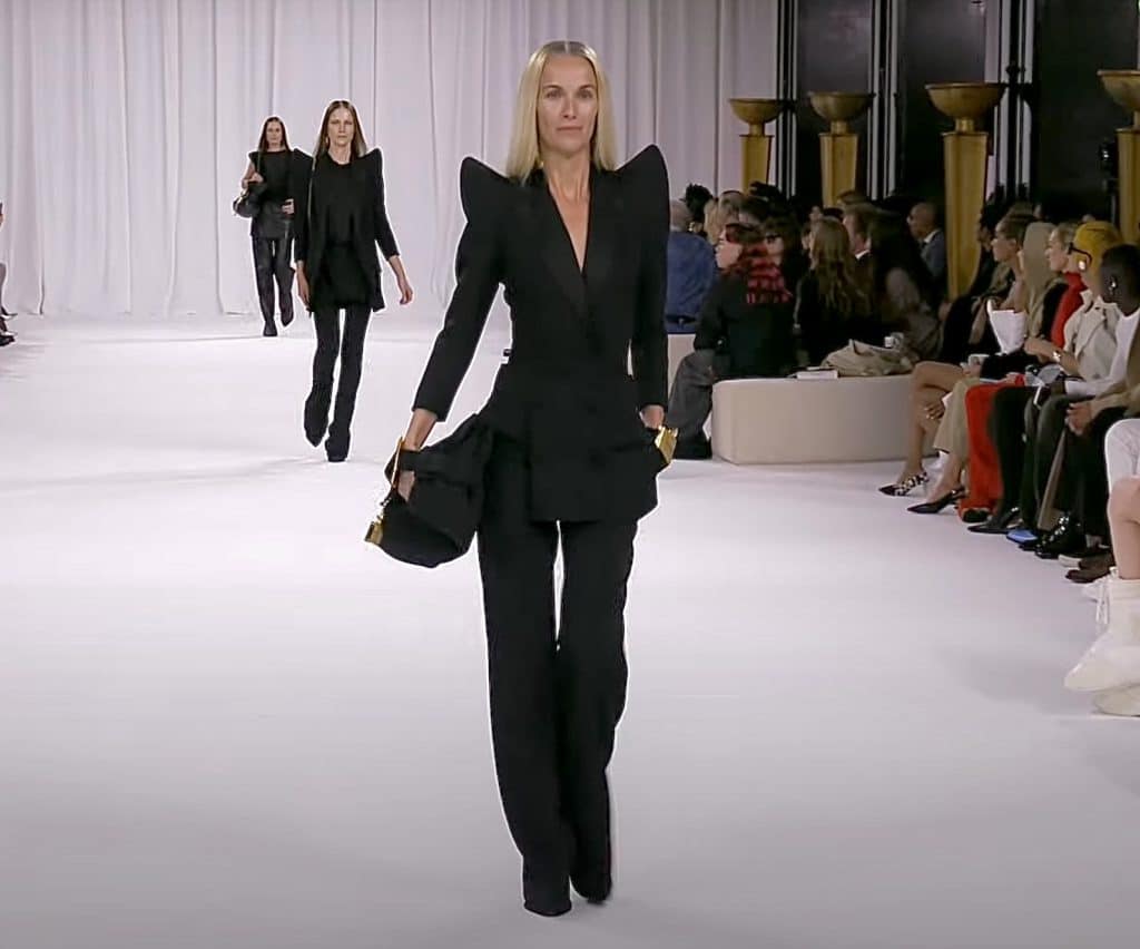 model walking a runway wearing an all black pantsuit outfit with shoulder pads